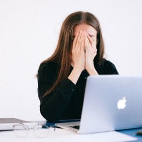 Vrouw met haar handen voor haar gezicht achter een laptop, overweldigd door werkdruk en constante bereikbaarheid