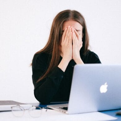 Vrouw met haar handen voor haar gezicht achter een laptop, overweldigd door werkdruk en constante bereikbaarheid
