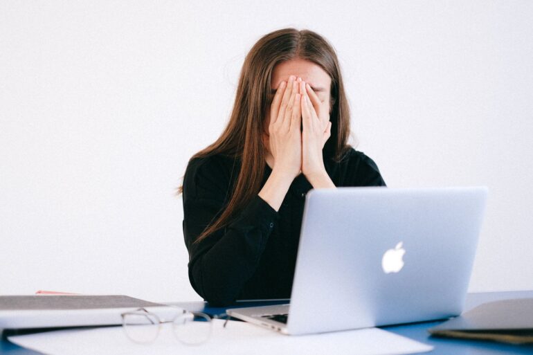 Vrouw met haar handen voor haar gezicht achter een laptop, overweldigd door werkdruk en constante bereikbaarheid